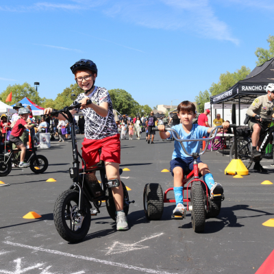 Children enjoying a biking activity at a busy outdoor event with crowds and tents in the background.