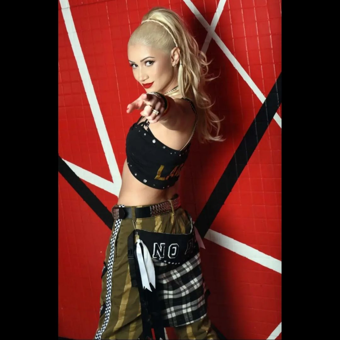 Blonde Pony-tailed Woman in performance-style clothing pointing toward the camera in front of a red geometric wall.