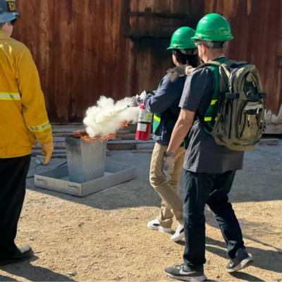 Promotional graphic for community safety training. Text reads: ‘CERT Fire Extinguisher Training.’ Image shows three individuals wearing green hard hats and reflective safety vests participating in a hands-on fire safety demonstration. One individual is using a red fire extinguisher to put out a small controlled flame in a metal container, producing white smoke. Another individual stands close by, observing and offering support, while a third individual wearing a yellow protective jacket oversees the exercis