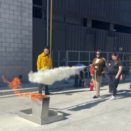 Fire personnel instructing two individuals how to use a fire extinguisher on a simulated trash can fire