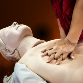 “Hands‑Only CPR Training.” Image shows an individual wearing a red top performing chest compressions on a medical training mannequin. The scene is tightly focused on the mannequin’s torso and the individual’s hands pressing down in the center of the chest. Background is softly blurred with warm tones suggesting an indoor instructional setting. Event labeled as public safety and health education.