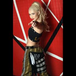 Blonde Pony-tailed Woman in performance-style clothing pointing toward the camera in front of a red geometric wall.